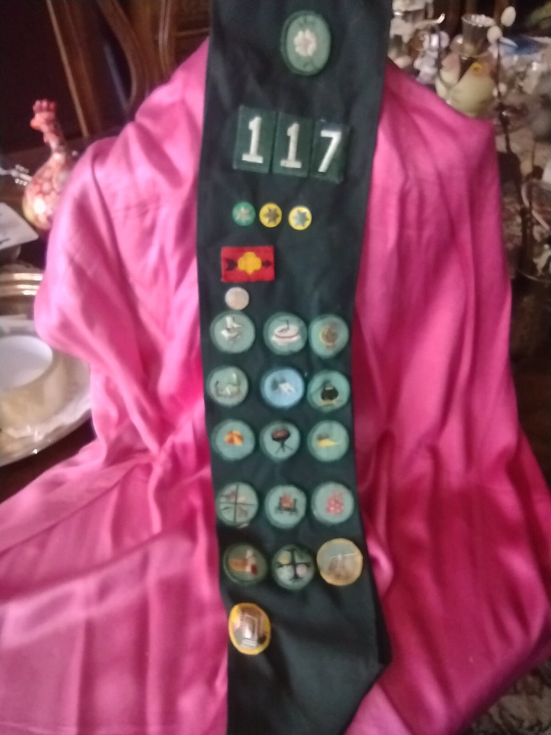 May include: A green sash with a number "117" and various badges and patches. The sash is displayed on a pink fabric background.