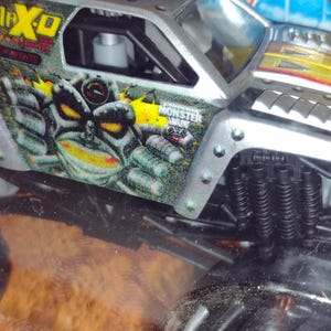 May include: A silver toy monster truck with black wheels and a green, yellow and black monster face graphic. The truck is labelled "Monster Jam" and "Max-D".