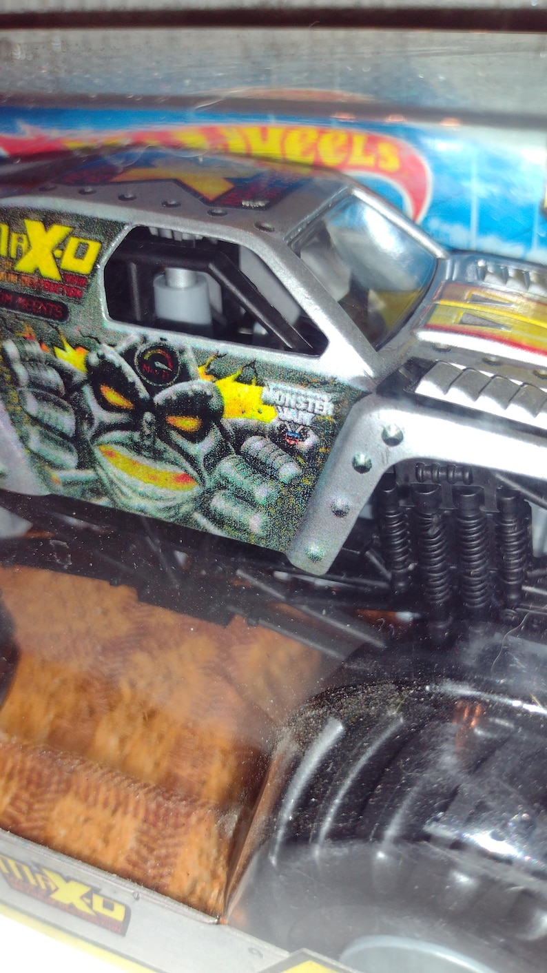 May include: A silver toy monster truck with black wheels and a green, yellow and black monster face graphic. The truck is labelled "Monster Jam" and "Max-D".