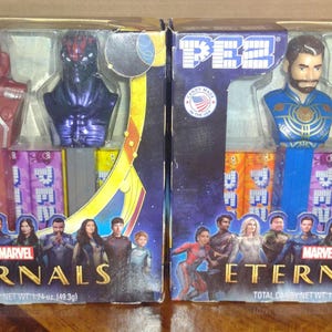 Puede incluir: Dos dispensadores de caramelos PEZ con personajes de Marvel's Eternals, empaquetados en cajas. Una caja tiene personajes rojos y morados, la otra tiene personajes azules y verdes. Las cajas muestran el logotipo de Marvel Eternals.