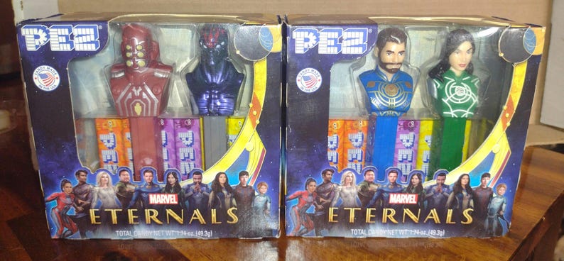 Puede incluir: Dos dispensadores de caramelos PEZ con personajes de Marvel's Eternals, empaquetados en cajas. Una caja tiene personajes rojos y morados, la otra tiene personajes azules y verdes. Las cajas muestran el logotipo de Marvel Eternals.