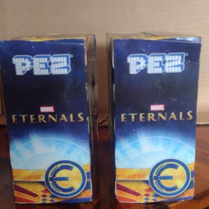 Puede incluir: Dos dispensadores de caramelos PEZ con el logotipo de Marvel Eternals. Las cajas son de color azul oscuro con texto blanco y un emblema circular. El logotipo de PEZ se muestra en la parte superior. Sobre una superficie de madera.