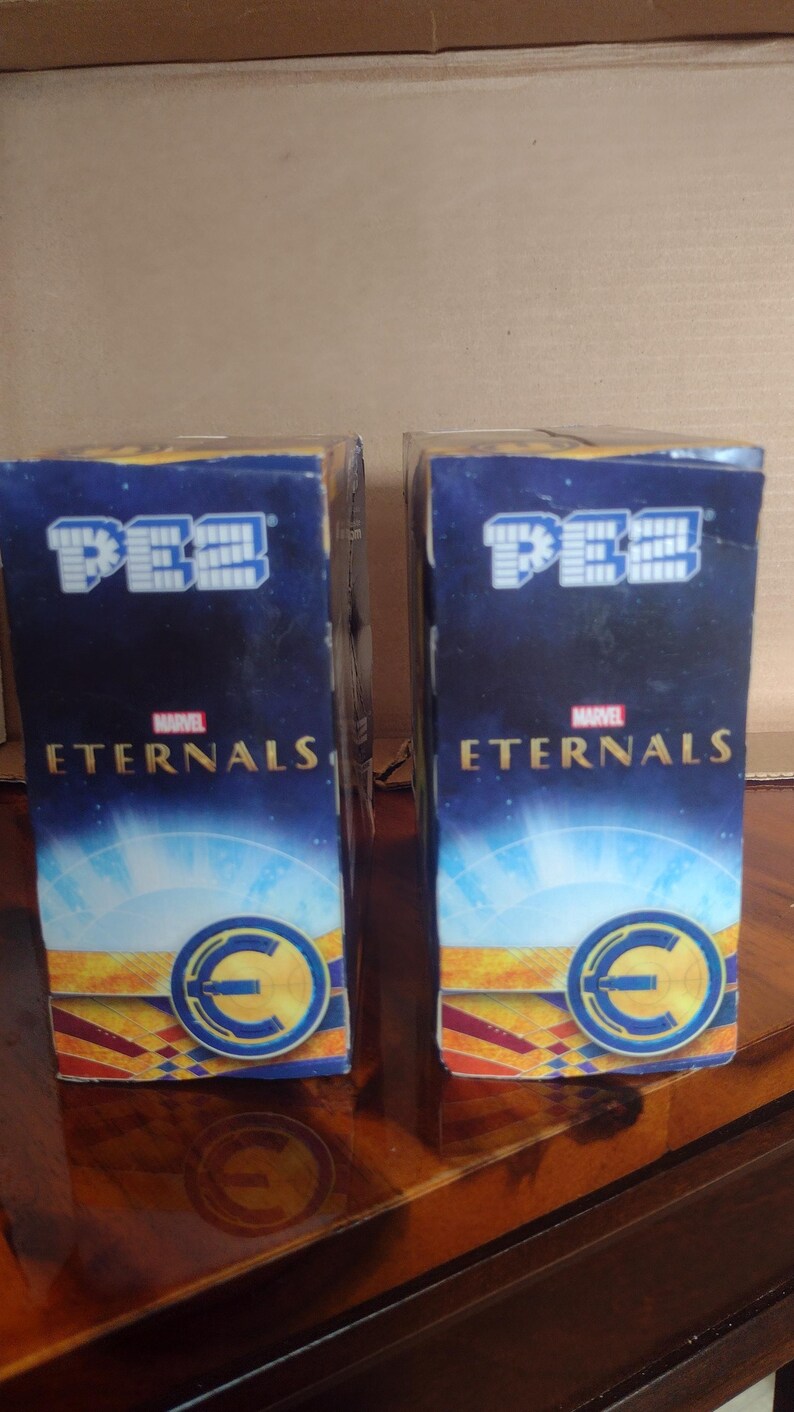 Puede incluir: Dos dispensadores de caramelos PEZ con el logotipo de Marvel Eternals. Las cajas son de color azul oscuro con texto blanco y un emblema circular. El logotipo de PEZ se muestra en la parte superior. Sobre una superficie de madera.