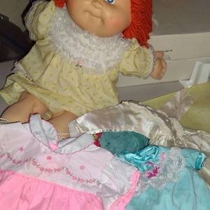 May include: A vintage Cabbage Patch Kids doll with red yarn hair and blue eyes, wearing a yellow dress with white lace trim. Several doll dresses in pink, white, and blue are also visible, suggesting a collection of doll clothing.