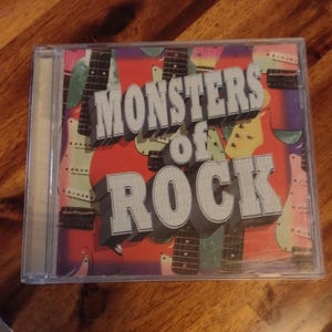 May include: A CD case with the title "Monsters of Rock" in a 3D, silver, block letter font. The background is red with a collage of guitar images in various colors. The CD case is clear plastic.