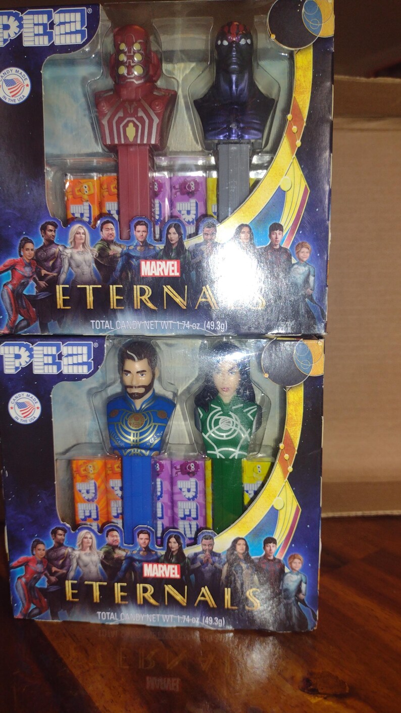 Puede incluir: Dos dispensadores de PEZ con personajes de Eternals de Marvel. El dispensador superior tiene un personaje rojo y dorado y un personaje p&uacute;rpura. El dispensador inferior tiene un personaje azul y dorado y un personaje verde. Cada caja tiene el logo de Marvel Eternals y el texto "PESO NETO TOTAL DE CARAMELOS 49,3 g".