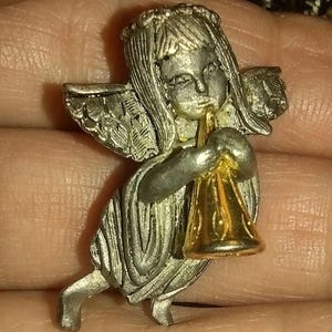 May include: A silver-toned angel pin with a gold-colored trumpet. The angel has detailed wings and a halo. The pin is held in a hand, showing the intricate design and craftsmanship of the jewelry.