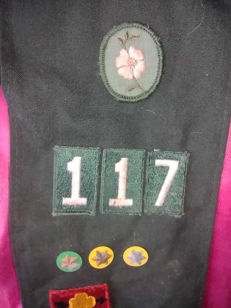 May include: A green fabric patch with the number 117 in white letters. There is a green and pink flower patch above the number patch. There are three small round patches with stars below the number patch.
