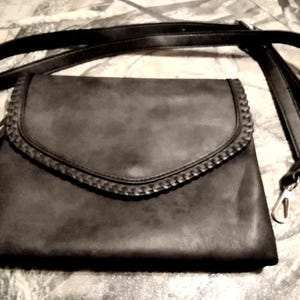 May include: A dark gray crossbody bag with a flap closure and a braided trim. The bag has a long, adjustable strap and a tassel detail. The hardware is silver-toned.