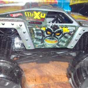 May include: A silver toy monster truck with black wheels and a Max-D Monster graphic on the side. The truck has a monster face with yellow eyes and teeth.