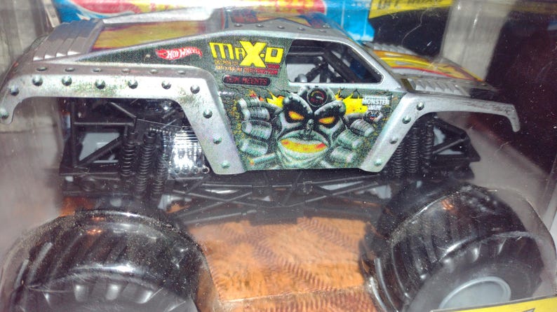May include: A silver toy monster truck with black wheels and a Max-D Monster graphic on the side. The truck has a monster face with yellow eyes and teeth.
