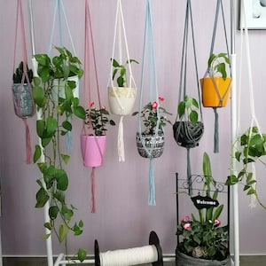 Può includere: Sei portavasi sospesi in macramè con cordini di diversi colori e anelli di legno appesi a un rack di metallo bianco. I portavasi contengono diverse piante in vaso, tra cui una vite verde, una pianta fiorita rosa e una pianta verde con fiori bianchi. Un rotolo di cordoncino bianco si trova sullo scaffale inferiore del rack.