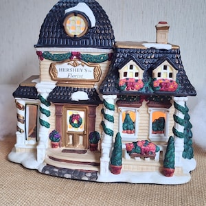 May include: A ceramic Hershey's Florist building figurine featuring a dark shingled roof with white snow accents, cream-colored siding, green garland trim, and red floral decorations. The front displays a sign reading HERSHEY'S Florist.