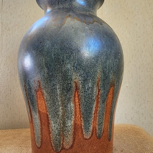 May include: A large, handcrafted ceramic vase with a wide body and a narrow neck. The vase features a blue-green glaze that drips down to reveal a rusty orange color at the base. The vase is approximately 18 inches tall.