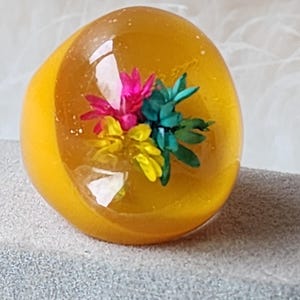 May include: A chunky, resin ring in a warm yellow hue, encasing a vibrant floral arrangement. The flowers are a mix of pink, yellow, and teal, creating a colorful contrast within the transparent resin. The ring is a bold statement piece.