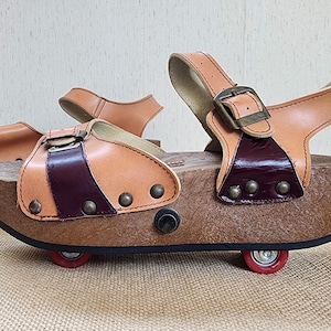 May include: A pair of vintage roller skates with a wooden platform and tan leather straps. The skates have a brown base with red wheels and metal buckles. The leather straps are accented with a darker burgundy stripe and metal studs.