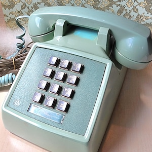 May include: A vintage mint green telephone with a square keypad featuring number and letter keys. The handset rests on top, and a coiled cord extends from the base. The phone has a retro aesthetic.