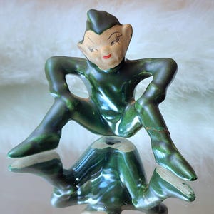 May include: A ceramic elf figurine in a dark green glossy finish, depicted in a crouching pose with hands on hips. The elf has a light-colored face with painted facial features, including red lips and dark eyes, and a pointed hat.