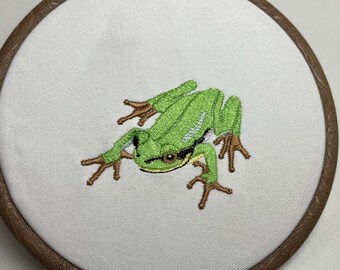 May include: Embroidered green frog on a white fabric background, framed in a brown wooden hoop.