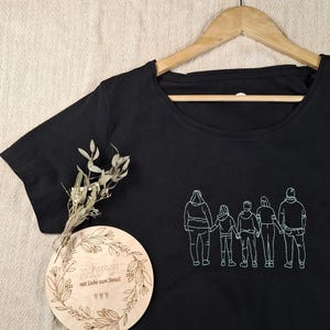 Pu&ograve; includere: Una t-shirt nera con un ricamo a linee bianche raffigurante una famiglia di cinque persone in fila che si tengono per mano. &Egrave; inclusa una targa rotonda in legno con il testo inciso "zauberhaft mit Liebe zum Detail" e un piccolo rametto di fiori secchi.