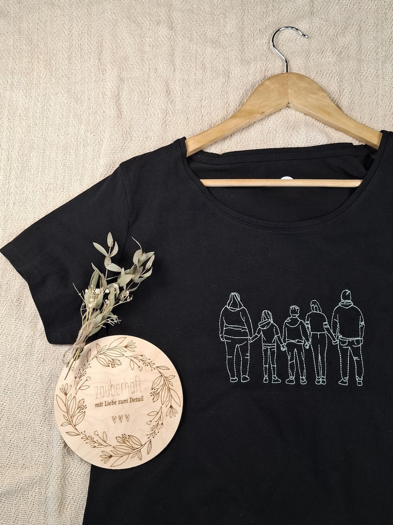 Pu&ograve; includere: Una t-shirt nera con un ricamo a linee bianche raffigurante una famiglia di cinque persone in fila che si tengono per mano. &Egrave; inclusa una targa rotonda in legno con il testo inciso "zauberhaft mit Liebe zum Detail" e un piccolo rametto di fiori secchi.