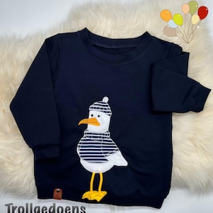 Pu&ograve; includere: Una felpa blu navy a maniche lunghe con un'applicazione centrale raffigurante un gabbiano bianco che indossa un berretto e un maglione a righe blu navy e bianche. Il gabbiano ha un becco arancione brillante e zampe palmate gialle. Testo Trollgedoens.