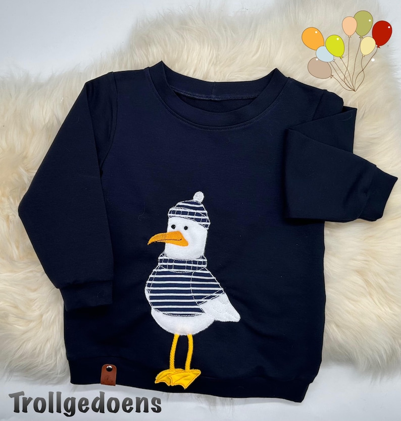 Pu&ograve; includere: Una felpa blu navy a maniche lunghe con un'applicazione centrale raffigurante un gabbiano bianco che indossa un berretto e un maglione a righe blu navy e bianche. Il gabbiano ha un becco arancione brillante e zampe palmate gialle. Testo Trollgedoens.