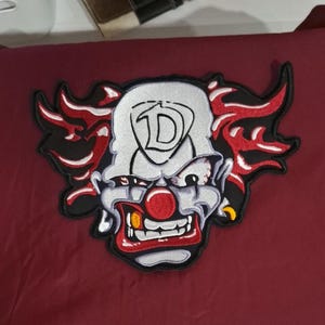 Pu&ograve; includere: Una toppa ricamata raffigurante un volto di clown con fronte bianca, naso rosso e un ampio sorriso con denti in vista. Il clown presenta fiamme rosse e nere stilizzate che si estendono dai lati della testa. Una grande lettera D &egrave; centrata sulla fronte.