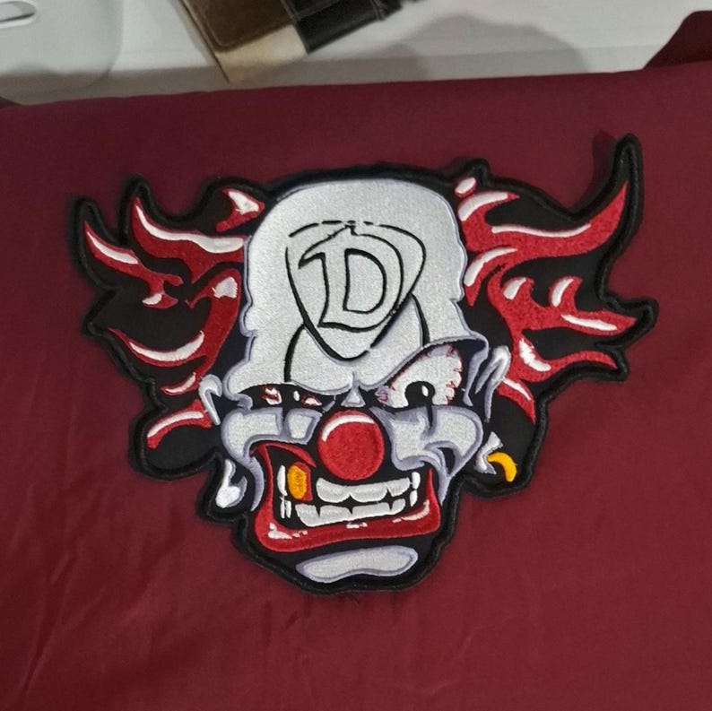 Pu&ograve; includere: Una toppa ricamata raffigurante un volto di clown con fronte bianca, naso rosso e un ampio sorriso con denti in vista. Il clown presenta fiamme rosse e nere stilizzate che si estendono dai lati della testa. Una grande lettera D &egrave; centrata sulla fronte.
