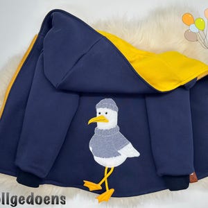 Pu&ograve; includere: Una giacca con cappuccio blu navy con fodera interna giallo brillante e cerniera gialla. Il retro presenta un grafico ricamato di un gabbiano bianco che indossa un maglione lavorato a maglia grigio e un berretto grigio, su zampe palmate.
