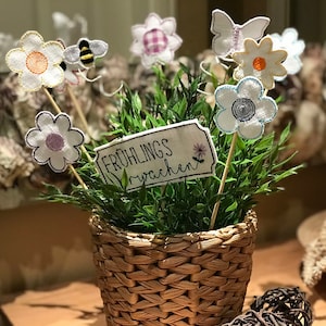 May include: A small potted plant with a woven basket, decorated with embroidered flowers, a bee, a butterfly, and a sign that reads "Frühlingswachen".