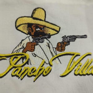 Pu&ograve; includere: Una toppa ricamata su tessuto bianco raffigurante un personaggio dei cartoni animati con un sombrero giallo che impugna due revolver grigi. Sotto il personaggio, il testo Pancho Villa &egrave; ricamato in corsivo giallo con contorno nero.