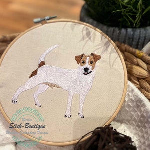 Könnte beinhalten: Stickerei eines weißen Jack Russell Terriers mit braunen Markierungen auf einem beigefarbenen Stoffhintergrund. Das Design befindet sich in einem runden Stickrahmen.