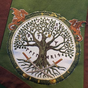 Pu&ograve; includere: Un pannello in tessuto verde con una toppa circolare ricamata. La toppa mostra un albero centrale con foglie verdi, due spade incrociate alla base e due draghi arancioni ai lati. Delle rune sono cucite sullo sfondo di colore bianco.