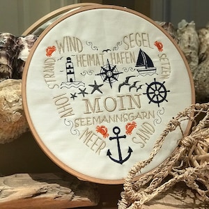 Puede incluir: Un aro de bordado blanco con temática náutica. El aro presenta un ancla, una brújula, un faro y las palabras "Wind, Strand, Ahoi, Heimat Hafen, Segel Setzen, Seemannsgarn, Meer, Sand, Moin".