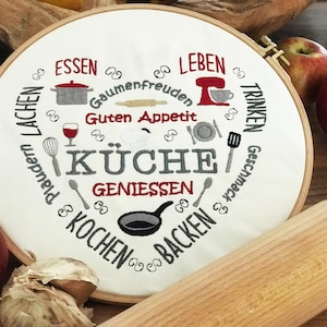 Puede incluir: Tela blanca bordada con un diseño en forma de corazón que presenta las palabras "Küche", "Essen", "Leben", "Trinken", "Kochen", "Backen", "Plaudern", "Lachen", "Geniessen", "Guten Appetit" y "Geschmack". La tela está estirada sobre un aro de bordado de madera. También hay manzanas y un rodillo en la imagen.