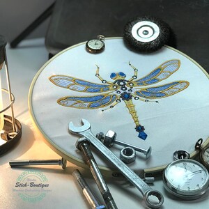May include: A white embroidery hoop with a blue and gold dragonfly design. The dragonfly is made of intricate details and has a steampunk aesthetic. The hoop is surrounded by tools and a pocket watch.