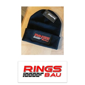 Pu&ograve; includere: Un berretto in maglia nero con risvolto e una toppa rettangolare. La toppa mostra la scritta RINGS BAU in rosso e bianco con una grafica a mattoni neri. Un'etichetta nera del marchio Beechfield &egrave; attaccata alla parte superiore del berretto.