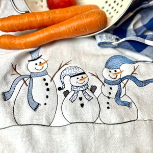 Puede incluir: Una toalla de cocina blanca con tres muñecos de nieve bordados. Los muñecos de nieve llevan sombreros y bufandas azules. Los muñecos de nieve sostienen palos en sus manos.
