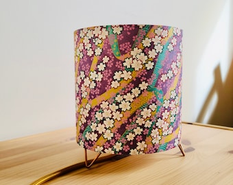 Puede incluir: Una lámpara de mesa con una pantalla de lámpara con estampado floral. La pantalla presenta un estampado floral rosa, blanco y verde con detalles dorados. La lámpara tiene un cable dorado y una base de metal dorado con tres patas.