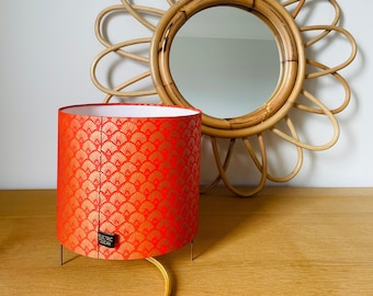 Puede incluir: Una pantalla de lámpara roja y dorada con un interior blanco se encuentra sobre una mesa de madera. La pantalla está sobre un fino soporte metálico de tres patas. Un espejo redondo tejido con un diseño de sol se encuentra detrás de la lámpara.