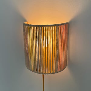 Op de afbeelding: Cilindrische wandlamp met een warme gloed. De kap is gemaakt van verticale strengen crèmekleurig touw, geaccentueerd met banden in geel, oranje en roze. De lamp is gemonteerd op een witte muur, met een gele stroomkabel.