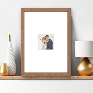 May include: A rectangular picture frame with a medium-brown wood grain finish and a wide white mat. The frame contains a centered photograph of a person in a white wedding dress and a person in a dark blue suit standing forehead to forehead.