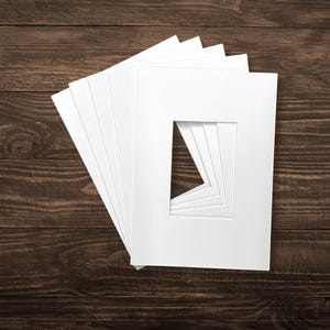 May include: A stack of five rectangular white paper photo mounts, each featuring a central rectangular aperture. The mounts are arranged in a fan-like formation, showcasing the clean, smooth texture of the white card material and the precise, sharp edges of the inner window openings.