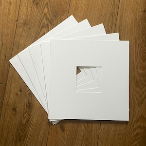 May include: A stack of five square white paper mounts, each featuring a central square aperture. The mounts are arranged in a fanned-out formation, revealing the layered edges of the square openings through the centre of the white cardstock stack.
