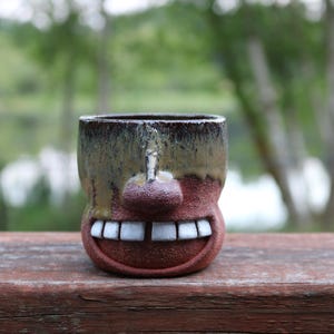 May include: A handmade ceramic mug with a face design. The mug features a brown and tan glaze, a nose, and a wide, smiling mouth with white teeth. The mug is set against a blurred outdoor background.