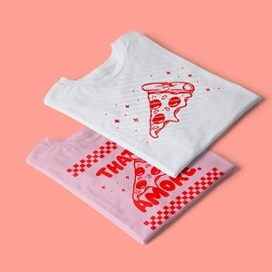Op de afbeelding: Twee opgevouwen t-shirts, één wit en één roze, met rode pizzapunt afbeeldingen. Het witte shirt heeft een pizzapunt ontwerp met sterren. Het roze shirt zegt "THAT AMORE" met een pizzapunt afbeelding en geblokte randen.