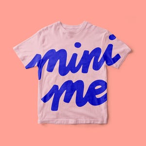 May include: A light pink short-sleeved t-shirt featuring the text mini me printed in a large, bold, blue cursive font across the front. The garment has a crew neckline and a standard fit, with the blue lettering extending across the chest area.