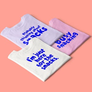 May include: Three t-shirts in white, lavender, and pink, each with a blue text design. The white shirt reads "I'm just here for the snacks." The lavender shirt says "probably thinking about snacks." The pink shirt says "busy snacking."