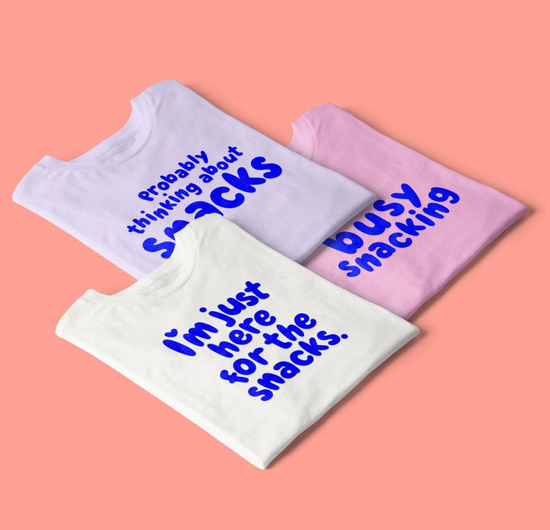 May include: Three t-shirts in white, lavender, and pink, each with a blue text design. The white shirt reads "I'm just here for the snacks." The lavender shirt says "probably thinking about snacks." The pink shirt says "busy snacking."
